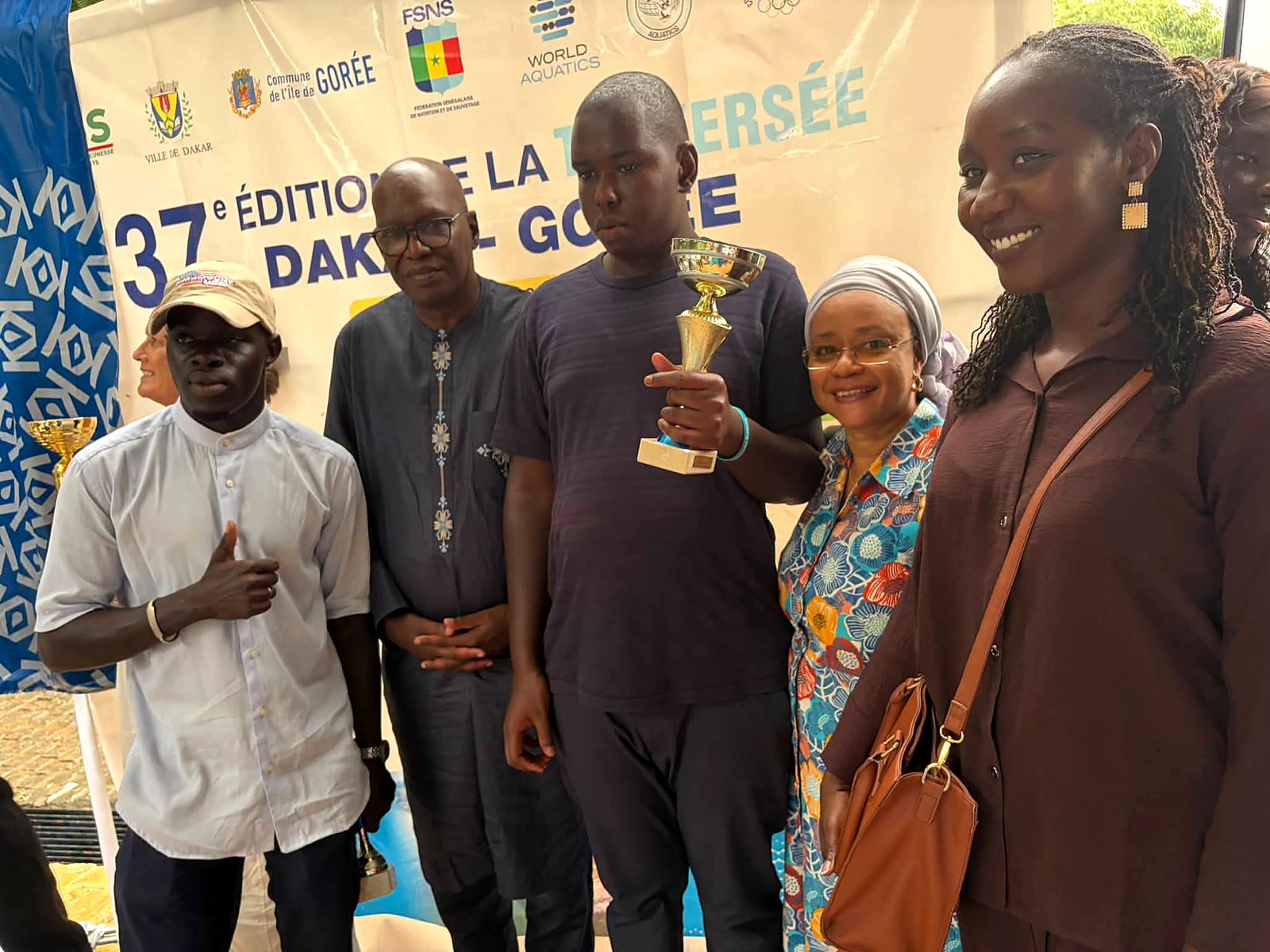 Mansour brandit son trophée, entouré de proches souriants devant la banderole de la Traversée Dakar–Gorée